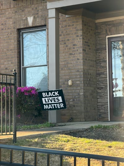 A "Black Lives Matter" sign stands near a brick-walled house in an outdoor setting. The house features windows with visible reflections and a light fixture on the brick wall. A small bush with pink flowers grows near the sign. A metal fence is partially visible in the foreground. Grass and greenery are present on the right side of the frame. The location is presumed to be in a residential area within the city of Atlanta, United States of America, based on visual cues such as architectural style and landscape. There are no people present in the image.