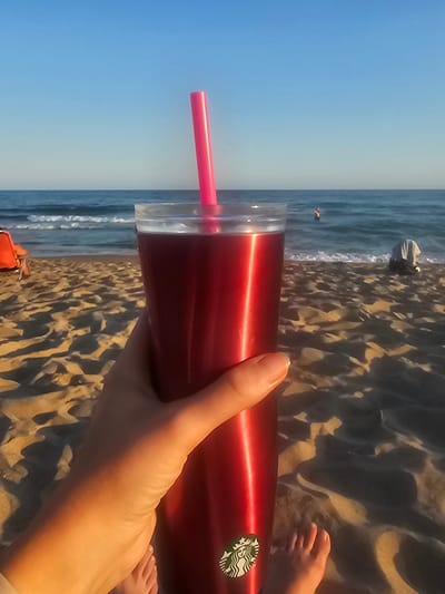 This is a vertically oriented, eye-level, first-person perspective medium shot of a person holding a red reusable tumbler with a pink straw on a sandy beach. The tumbler has a Starbucks logo on it. The person's hand is visible holding the tumbler, and their feet are also visible in the foreground. The background consists of the ocean and a clear blue sky, suggesting a sunny day. The lighting indicates it is likely late afternoon or early evening, given the long shadows cast on the sand. The scene appears to be a relaxing vacation or outing at the beach.