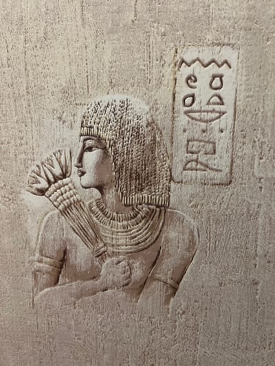 This media file shows a close-up of an ancient Egyptian-style relief carved into a rough, textured surface, possibly stone or plaster. The relief depicts a profile of a person, likely a woman, with distinct features. The figure wears an elaborate headdress or hairstyle with multiple layers and decorative elements. A necklace or collar, also layered, is prominent around the neck. The figure's hands are clasped at the chest, with finely detailed fingers. To the left of the figure, there is a rectangular cartouche containing hieroglyphic symbols. These symbols appear to be a zigzag line, two circular shapes, a spiral, and a shape resembling a half-circle or an oval with a line. The carving style is detailed, with a clear distinction between raised and recessed areas, suggesting age and possibly wear. The lighting highlights the texture of the material and the relief work. The overall impression is one of historical or artistic significance, likely a decorative element from a tomb or temple. There is no information about the time of day, weather, or emotions conveyed, as the focus is on the static carving. The context of "Ladozhskoe Ozero, Russia" does not appear to be directly related to the visual content of the image itself, which strongly suggests an ancient Egyptian setting.