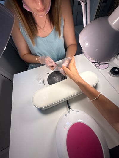 The image depicts a close-up view of a nail salon setting, likely in Simferopol, Ukraine. The scene is captured from a high-angle perspective, focusing on a nail technician and a client at a white table. The technician, wearing a light blue sleeveless top, transparent gloves, and a pink face mask, is positioned on the left. Her hands, also gloved, are holding a metal nail tool, actively working on the client's right hand. The client's arm, adorned with a delicate pearl bracelet, is resting on a white padded armrest positioned on the table. A pink-accented nail curing lamp is visible in the foreground, and a white nail dust collector with a circular fan is situated behind the armrest. Other items on the table include a small jar with a black lid, a pair of cuticle scissors, and what appears to be a nail file or pusher resting on a pink mat. The overall environment suggests a professional nail care service is in progress.