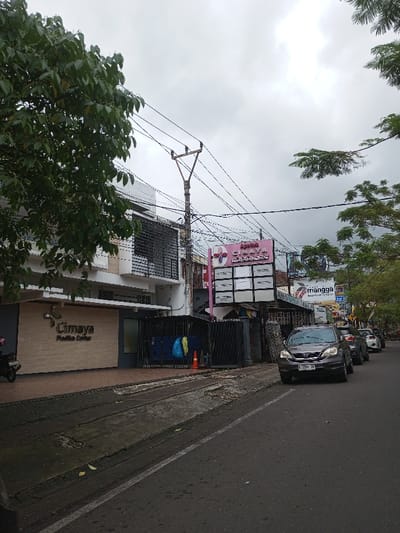 The image depicts a street scene in Vaatharawattai, Sri Lanka, under an overcast sky. On the left, a multi-story building with a light-colored facade displays "Cimaya Medika Center" in dark lettering. A dark scooter is parked partially on the sidewalk in front of this building. To the right of the first building, a large utility pole rises, laden with numerous overhead wires. Adjacent to the utility pole, a building features a prominent pink sign with "Apri Cimaya" and a cross symbol, below which are multiple white rectangular panels. Dark metal gates are positioned in front of this building, behind which blue and yellow items are visible, alongside an orange traffic cone. The asphalt road occupies the foreground and extends into the distance on the right, where a dark gray Honda CR-V (license plate E 2304 JF) is either parked or moving slowly, followed by several other parked vehicles. A concrete sidewalk or drainage channel runs parallel to the road on the left. Green trees with dense foliage are visible on the far left, partially obscuring the upper portion of the first building, and further down the street on the right. No individuals are discernible in the scene.