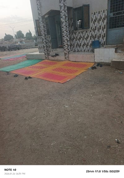 A modest building with patterned tile walls sits on a packed-earth surface. Multiple prayer rugs in varying colors, including red, yellow, and green, are spread in front of the building. Several pairs of shoes are neatly arranged near the rugs and entrance, indicating a place of prayer or gathering. A few utility vehicles are parked in the distance.