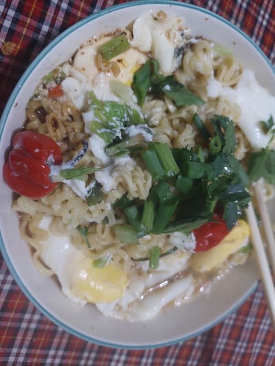A bowl of instant noodles with toppings including chopped scallions, cilantro, a fried egg, and what appear to be small red peppers sits on a red, black, and white checkered tablecloth. The scene is likely in a home kitchen or dining area in Jelutung, Indonesia. There are no people visible in the image.