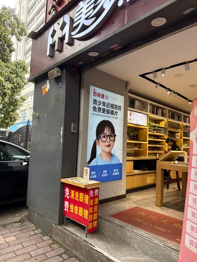 In Yuexiu District, China, a storefront for an optical shop is visible. The shop features large, illuminated signage with Chinese characters. A prominent advertisement on the left side of the entrance showcases a young girl wearing glasses, promoting a campaign for children's myopia prevention and free eyeglasses replacement. Below this advertisement, a red stand displays smaller signs offering services such as free eyeglass cleaning and repair. The interior of the shop, seen through the open entrance, reveals shelves stocked with various eyeglasses. A woman is observed inside, seemingly attending to a customer or organizing the merchandise. A red welcome mat with Chinese characters and the English word "Welcome" is placed on the floor at the entrance. The street outside the shop is paved with bricks, and a black car is parked to the left. The building to the left of the optical shop is a multi-story structure with numerous windows.