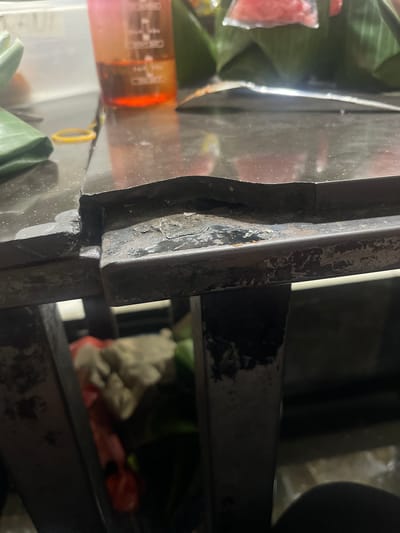 The image shows a close-up view of a dark, reflective surface, possibly a table or countertop, with a visible seam in the middle that shows signs of wear and tear. The surface appears to be made of a dark material, possibly laminate or stone, with some scuff marks and a slight warping in the area of the seam. To the left, a green object, perhaps a piece of fabric or plastic, is partially visible, along with a yellow rubber band. In the background, a partially filled plastic container with a pink and orange gradient holds a reddish liquid. Further back, out of focus, are some objects wrapped in banana leaves, suggesting food items. The scene appears to be in an indoor setting, with a dimly lit environment and some reflections on the surface. There are no people visible in the image. The location in Barat Yaman, Indonesia, is not explicitly depicted or suggested by any visual cues in the image itself.