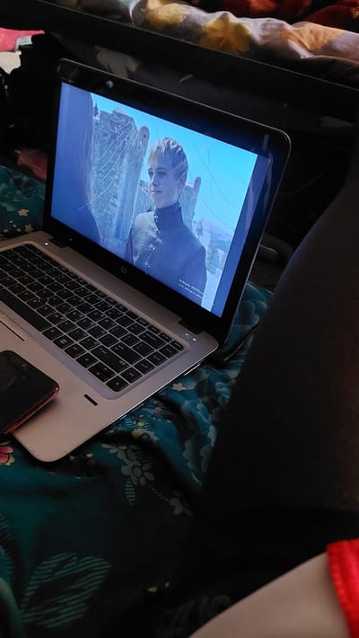 The image shows a laptop with a cracked screen displaying a scene from a show, likely Game of Thrones, featuring a young blonde character. The laptop is open and placed on a patterned fabric, possibly a blanket or bedspread, with floral designs in shades of teal and yellow. A red smartphone is visible on the left side of the laptop. The background appears to be a room, with a dark object, possibly a bag or furniture, on the top right. The lighting suggests an indoor environment. The image's context of New Nyanya, Nigeria, is not directly evident from the visual elements.