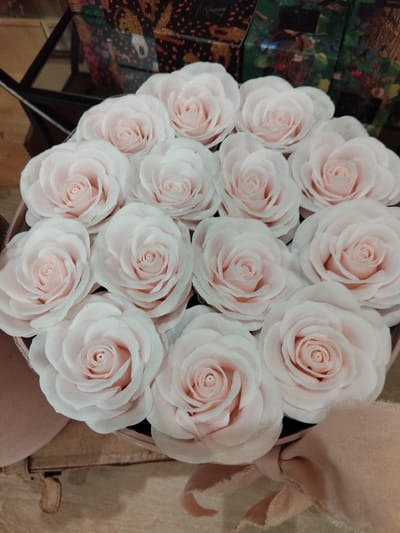 This is a close-up, overhead shot of a bouquet of pale pink, rose-like flowers, possibly camellias, arranged in a rounded arrangement within a decorative box. The flowers are tightly packed, with multiple layers of soft, delicate petals forming distinct centers. The lighting is soft and diffused, suggesting an indoor setting, possibly a flower shop or a home.
In the background, to the left, there's a glimpse of a dark, patterned surface with intricate designs in shades of green, black, and orange, possibly a decorative box or fabric. To the right and behind the flowers, a piece of pale pink fabric with a slightly frayed edge is visible, adding a soft, textural element. The overall impression is one of elegance and softness, with the delicate colors and textures creating a serene atmosphere.
No people are visible in the image. The focus is entirely on the floral arrangement. There are no clear indicators of time of day or specific weather conditions. The location context of Vila Nova de Famalicão, Portugal, does not offer any direct visual cues within this specific image. There is no visible text in the media file.