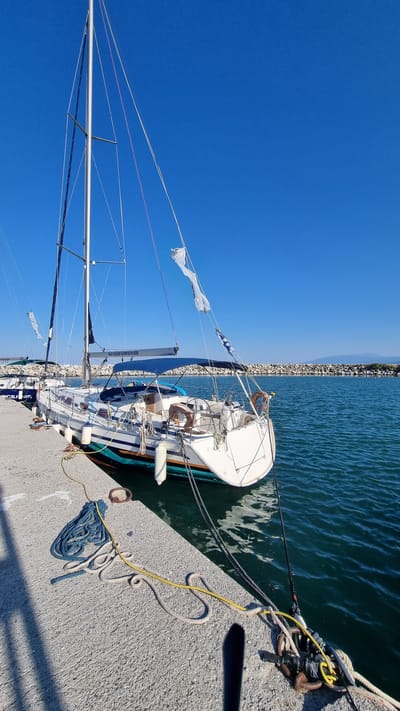 The image depicts a sailboat docked at a pier in Limani Litochorou, Greece. The sailboat, a Bavaria Cruiser, is white with teal and orange accents along its waterline. It is moored with thick ropes to the concrete pier. The boat has a tall mast with several lines running to the deck, and a blue canopy is extended over the cockpit. Two white fenders hang from the port side of the hull. Several white flags are attached to the mast, two of which appear tattered and are flapping in the breeze. A second sailboat is visible further down the pier.
The pier is constructed of concrete and shows signs of wear. Various ropes, including a coiled blue rope and a yellow rope, are scattered on the pier's surface. A rusty metal ring is visible near the coiled blue rope. The water is a clear, deep blue-green and appears calm with small ripples.
In the background, a breakwater made of large stones separates the harbor from the open sea. Beyond the breakwater, a low, hilly coastline is visible under a bright, clear blue sky. The sun casts shadows on the pier, indicating it is daytime. No people are visible in the image.