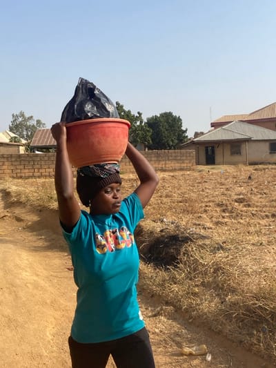 An adult Black female is depicted walking on a dirt path in Udu, Nigeria, carrying a large red plastic basin containing a black plastic bag balanced on her head. Her arms are raised, with hands holding the sides of the basin for stability. She wears a teal short-sleeved t-shirt with a multicolored graphic, black trousers, a brown knit cap, and a patterned fabric wrap beneath the basin. A small white cross-shaped earring is visible on her left ear. Her gaze is directed towards the right side of the frame. The immediate environment features a light brown dirt path bordered by dry, golden-brown grasses and sparse vegetation. In the background, a low brick wall separates the foreground from several buildings, including one with a grey corrugated metal roof and a dark door, and another with an orange-brown tiled or metal roof. Green foliage from trees is visible behind the wall and buildings. The sky above is clear and light blue.