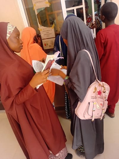 The scene is set inside a public service or registration office in Kwazailewa, Nigeria. Six individuals are present. In the foreground, a young female with light brown skin, wearing a maroon hijab and dress with a patterned forehead band, holds an open document or book in her left hand and a pink-cased smartphone in her right. She directs her gaze towards the right. To her right, another young female, partially viewed from behind, wears a dark grey hijab and dress, with a pink backpack adorned with multiple charms slung over her right shoulder. Her right hand is extended towards the documents held by the first female, suggesting an interaction or exchange. Both foreground females wear flat footwear; the one in grey has visible patterned anklewear.
Behind a white-framed glass partition, three individuals are visible. One person, largely obscured, wears an orange head covering and holds a phone to their ear. To their right, a young male in a red long-sleeved shirt is partially visible. Further right, another male in a patterned shirt (red, white, black) is visible through the glass. A white rectangular sign is taped to the glass partition, displaying "VOTER PLASTIC ID CARD IS AVAILABLE" and the number "8067503370", along with a small red, green, and white segmented graphic. The background includes a light-colored wall.