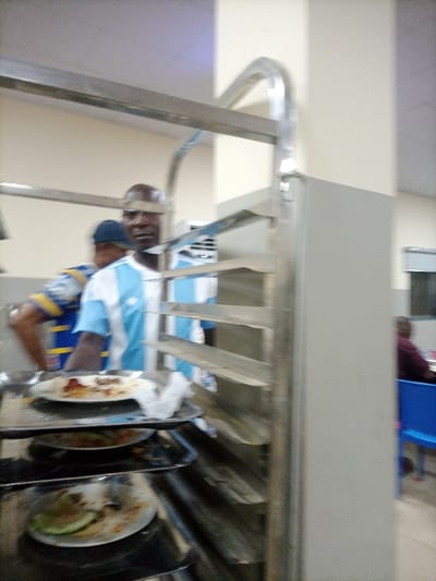 A man with a bald head is seen behind a metal dish rack. He's wearing a white and light blue striped jersey. Plates with leftover food are stacked on the rack. In the background, another person is wearing a blue and yellow shirt. A person sitting on a blue chair is visible to the right. The setting appears to be a cafeteria or similar dining area.