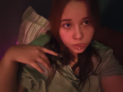 A young person with long brown hair is lying down, propped up by a pillow with stripes. They are wearing a green satin top. The person is looking to their right with a neutral expression. A dim, warm light illuminates the scene, casting shadows. The background is dark. The image is grainy. This scene is not identifiable as being in Ekibastuz, Kazakhstan.
