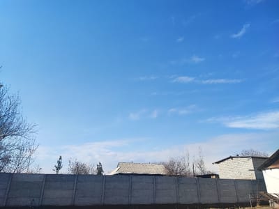 The image depicts an outdoor scene in Khidirsho, Uzbekistan, under a clear blue sky with scattered cirrus clouds. A long, grey concrete panel fence, composed of multiple rectangular sections, extends horizontally across the lower half of the frame. The ground directly in front of the fence is dark earth. Behind the fence, various bare deciduous trees and some evergreen trees are visible. Architectural structures include a building with a light-colored, pitched roof partially visible to the left of center, and a rectangular building constructed of light grey concrete blocks with a flat roof line to the right of center. A portion of another building with a dark, angled roof is visible on the far right. No individuals or discernible actions are present.