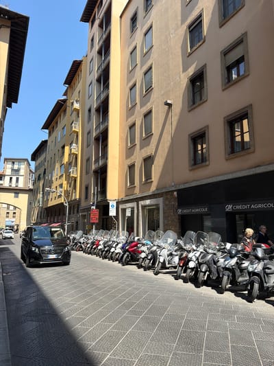 This is a street-level view of a sunlit narrow street in Florence, Italy, lined with tall, multi-story buildings. The buildings have distinct architectural features, including stucco facades in shades of beige and yellow, numerous windows with dark frames, and some balconies. The street is paved with patterned gray tiles. On the right side of the street, a row of parked scooters and motorcycles occupies a significant portion of the sidewalk and street. Some scooters are parked in front of the Crédit Agricole bank. Two individuals, a woman with curly blond hair and a man with dark hair, are visible near the scooters, with the woman looking towards the viewer. A black van is parked on the left side of the street, facing the camera. Further down the street, other vehicles and pedestrians are visible, and a large arched opening suggests another street or entrance. The sky is clear and blue. A red sign with white text is mounted on a pole on the right side of the street.