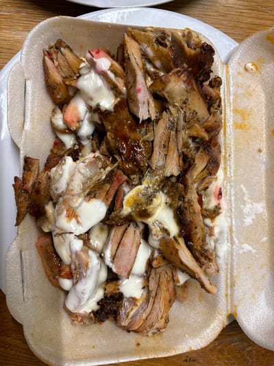 The image shows a close-up of sliced meat with white sauce in a white styrofoam container. The meat appears to be a mix of light and dark pieces, some glazed with a darker sauce. The container is rectangular with rounded corners and is sitting on a wooden surface. The location is Oswestry, United Kingdom. There are no people visible.