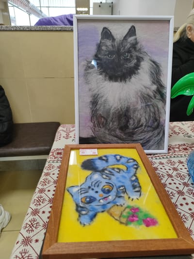 The image shows two framed cat artworks on a patterned tablecloth. The artwork in the foreground depicts a blue cat with stripes against a yellow background, framed in wood. The artwork behind it is a portrait of a long-haired cat with black and white fur, framed in white. The items are on a table in what appears to be an indoor setting. The location, Fryazino, Russia, cannot be forensically determined from the image.