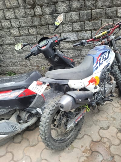 The media file shows a close-up, overhead view of two damaged motorcycles lying on a cobblestone and paver brick surface. One motorcycle is primarily white with blue and red graphics, including a Red Bull logo. It has a knobby tire, suggesting it's an off-road or dual-sport model. The other motorcycle is black and appears to be a scooter, with a digital display on the handlebars and a side stand. Both vehicles are tilted and appear to have fallen.
The setting is an outdoor area paved with textured stones and interlocking pavers. There are small patches of dry grass growing between the paving stones. The lighting suggests it is daytime. The overall impression is one of disarray and potential accident.
Visible text on the white motorcycle includes "PULSE" in large letters and what appears to be the word "TVS" in smaller print below it, along with some Hindi script. The license plate on the black scooter is partially visible and shows the characters "A789V". Text on the black scooter's bodywork is also visible, including "KAPOOR MOTORS".
The scene is static, with no people or animals present. The focus is solely on the damaged motorcycles and their immediate surroundings. The condition of the motorcycles and the way they are lying suggest they may have been involved in a crash.