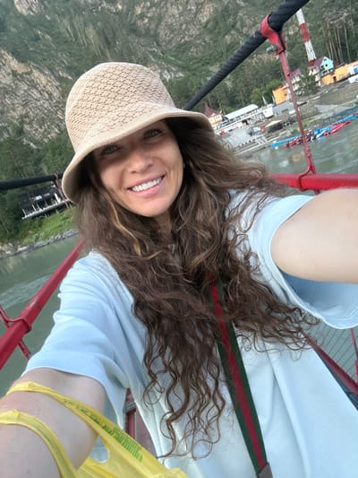 A woman wearing a light blue shirt and a straw hat takes a selfie from a red suspension bridge. She has long, curly brown hair and is smiling at the camera. The bridge spans a wide, green river, with boats docked on the far bank. In the background, a cluster of colorful buildings and a steep, tree-covered mountainside are visible. The scene appears to be in or near the city of Gorno-Altaysk, Russia, along the Katun River. The overall atmosphere is one of natural beauty and outdoor recreation.