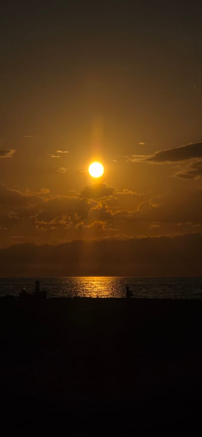 A vertical, full-frame shot captures a sunset over the sea at Boz Beshik, Kyrgyzstan. The sun, a bright, orb-like sphere with a luminous halo, hangs centrally in the sky, casting a golden path of light across the rippling water. Clouds, tinged with orange and gold from the setting sun, are scattered across the sky, with some appearing darker and more solid. In the foreground, silhouetted against the bright water and sky, are the dark shapes of people and possibly beach furniture on a sandy shore. The overall impression is one of tranquility and natural beauty.
