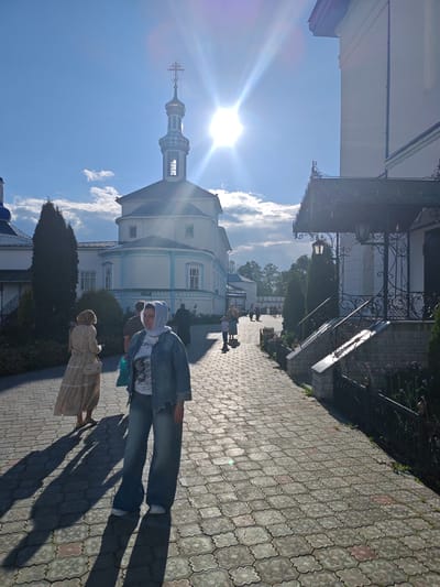 The image depicts a sunlit scene within the grounds of the Raifa Monastery in Raifa, Russia. The perspective is from a paved pathway, composed of interlocking gray and pink decorative stones. A woman in the foreground, wearing a light blue denim jacket, a white t-shirt with a graphic print, and wide-leg jeans, is facing the camera, looking slightly to her right. She has a light-colored hijab covering her head. To her left and further down the path, another woman in a long, light-colored patterned dress and a light-colored purse is walking away from the camera, casting a long shadow. Several other individuals are visible in the midground and background, appearing to walk along the pathway.
Dominating the background is a white, multi-story building with a prominent blue-domed roof and a tall, slender tower topped with a cross. The building features arched windows and decorative blue trim. To the right of the pathway, there is an entrance with a dark, ornate metal awning and a set of stone stairs with a similar metal railing. Cypress-like trees are strategically placed along the path and near the buildings. The sky is bright blue with scattered white clouds, and the sun is prominently visible, creating a strong glare and lens flare effect. The overall impression is of a serene, religious site bathed in bright sunlight.