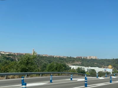 The image captures a wide-angle view of a road and a distant town under a clear blue sky. The foreground shows a paved road with white lane markings and several blue and white striped bollards lining the edge. A metal guardrail runs along the side of the road. Beyond the road, the landscape rises into a tree-covered hill.
On the hilltop, a town with numerous buildings is visible, characterized by terracotta roofs and some taller, modern apartment blocks. A prominent church tower with a spire stands out against the skyline, suggesting a historical or religious landmark within the town. Further to the right, a large industrial building with white walls is situated among the trees. A sign for "FELIU PACKAGING" is visible on this building, providing a specific location cue.
The scene appears to be set during the daytime, with bright sunlight indicating fair weather. The clear sky and the vibrant green of the trees suggest a pleasant, possibly summer day. The overall impression is one of a calm, rural or semi-rural setting with a developing urban area in the distance. The perspective suggests the photo was taken from a moving vehicle on the road, looking towards the hillside and town. There are no people visible in the image.