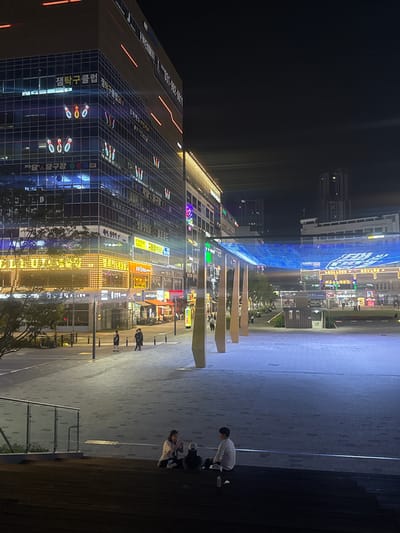 The image, oriented correctly from its original sideways capture, presents a lively urban night scene, likely in Gimpo-si, South Korea. On the left, a large multi-story commercial building dominates the frame, its glass facade reflecting the vibrant, colorful neon and LED signs of various businesses. These signs primarily display Korean text, including "챔탁구클럽" (Cham Table Tennis Club), "생탁구클럽24" (Saeng Table Tennis Club 24), and "당구장" (Billiards Hall), indicating entertainment venues. Other visible Korean phrases include "오락랜드" (Amusement Land), "계명진", "명품방질출", "헤이, 정착역", and "이스", along with numbers like "180".
Spanning above a pedestrian plaza in the mid-ground is a prominent blue light installation, creating an archway or canopy of glowing light. Below this, smaller trees are interspersed among more illuminated storefronts. The plaza floor is a mix of patterned gray pavers leading to a darker, possibly wooden or tiled, raised area or steps on the far right.
Several people are present in the scene. On the steps to the right, two individuals are seated, looking down, while another person in a light-colored top is bent over in the foreground, seemingly engaged with something on the ground. In the distance, other figures can be seen walking near the brightly lit shops.
The time of day is clearly night, with the sky a deep black and all illumination coming from artificial light sources. The atmosphere is vibrant and active, typical of an urban entertainment district after dark. The weather appears clear, with no signs of precipitation, allowing for good visibility of the detailed lighting and structures.