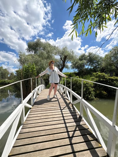 An outdoor scene is depicted from an unusual, rotated perspective, showing a woman on a wooden bridge under a bright sky. The woman, with blonde hair, is wearing a hat, sunglasses, a white long-sleeved shirt, black shorts, and white sneakers. She stands on the bridge with her arms outstretched, leaning back, appearing to balance or pose for the camera.
The bridge features dark wooden planks for the walkway and a white, angular railing on one side, extending diagonally across the frame. Shadows from the railing and the woman are sharply cast on the bridge planks, indicating direct sunlight and a clear day. To the right of the bridge, a body of water (likely a river or canal) is visible, reflecting parts of the sky and surrounding greenery. On the left side, lush green trees and dense foliage line the water's edge, creating a natural backdrop. Above and below the bridge, a blue sky dotted with scattered white clouds fills the background. No text is visible. The scene suggests a relaxed moment in a scenic natural environment, specifically Gay-Kodzor, Russia.