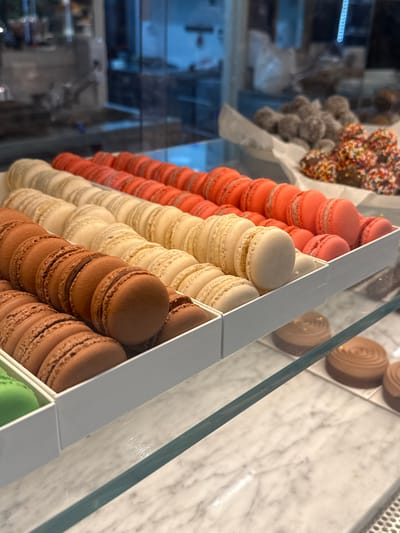 A bakery display case contains rows of macarons in various colors: orange, cream, and chocolate. Behind them are pastries covered in sprinkles. The display case has a marble surface and a glass top. The location is a bakery, possibly in Al Jizah, Jordan. No people are visible.