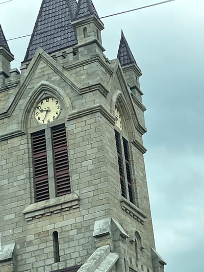 The image shows a close-up view of a stone clock tower, likely part of a church or public building, against a cloudy sky. The tower has a gothic architectural style, with pointed spires and arched windows. A clock face is visible on the tower, with Roman numerals and hands indicating the time. Below the clock are louvered windows. The stone construction is detailed, showing individual blocks and decorative elements. The scene is devoid of people or any immediate human activity. The location is identified as Oituz, Romania.