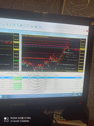 This is a medium shot, angled photo of a laptop screen displaying a trading platform. The laptop is black and has a Samsung logo visible on the bottom bezel. On the right side of the laptop, a white and colored remote control is resting. The screen shows a financial chart with candlestick patterns in red and green, indicating price fluctuations. Several labels and data points are visible on the chart, including "BTCUSD M1", price levels like "66348.540", "66155.160", and timestamps such as "--00:18". There is also a table on the right side of the screen with columns labeled "Цена", "S/L", "T/P", and "Прибыль", displaying numerical data and "Комментарий" with text like "Hybridfx-v2-0". The system clock in the bottom right corner of the screen shows "21:55" and the date "17.06.2026". The overall setting appears to be indoors, likely a home or office, given the context of a laptop and remote control. There are no people visible in the frame. The image has a red notification light illuminated on the laptop bezel.