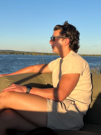 A man sits on an olive-green seat with a body of water and vegetation visible in the background, in Victoria Falls, Zimbabwe. He is wearing sunglasses, a light-colored t-shirt, and light-colored shorts with a watch on his left wrist. The man is seated with his legs crossed and is gazing downwards to his left. Sunlight illuminates the scene, creating shadows on the man and the seat. A steering wheel is partially visible to the right.