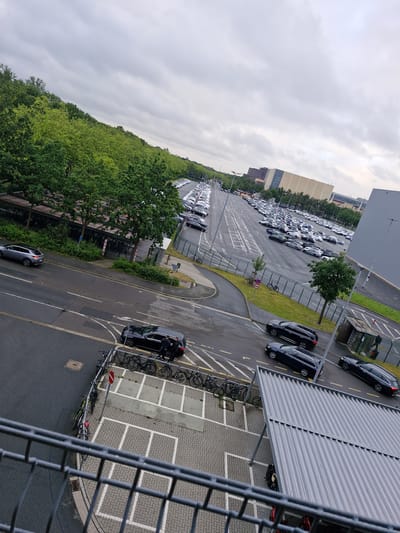 The image shows a large outdoor car park and a road, with a building and trees in the background. The car park is filled with a large number of white and dark-colored cars, arranged in rows. There are also some trees lining the edge of the road and the car park. In the foreground, there is a metal railing and a section of a building with a corrugated metal roof. The sky is overcast with clouds. Two dark-colored cars are parked on the road, and two individuals in dark clothing are standing next to one of them. Several bicycles are parked on a designated rack near the road. The scene appears to be a typical urban environment, likely in a car dealership or a large industrial area. The presence of many new cars in the lot suggests a car dealership or a distribution center. The location is identified as Bremen, Germany.
