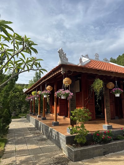 This is an exterior shot of a traditional building, potentially a temple, in Liên Nghĩa, Vietnam. The building has a red-tiled roof with ornate white trim. Hanging from the eaves are decorative lanterns and potted plants with pink and white flowers. Wooden pillars support the roof. A garden bed with red and green plants runs alongside the building. The foreground features a tree with large, green leaves and pale flowers. A stone path leads towards the building, and lush greenery surrounds it. The sky is blue with scattered clouds.