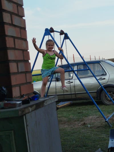A young girl with blonde hair is climbing on a blue metal swing set. She is wearing a green tank top with black trim and blue shorts with a white pattern. Her left hand is raised in a wave. She is wearing white and pink sandals. Behind her, a silver car is parked on a grassy area. The background shows a cloudy sky and a field of grass. To the left of the girl, a brick structure is visible, and on a wooden surface in the foreground, a blue screwdriver and other objects are present. The scene appears to be outdoors in a rural setting, possibly in Крывасельскі сельскі Савет, Belarus.
