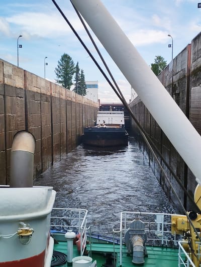 The image depicts a canal lock with a barge navigating through it. The lock walls are constructed from large, weathered wooden planks, with a prominent metal pipe extending from the left wall into the water. The water is turbulent, suggesting the lock is actively in operation. In the distance, another barge, this one white with a darker hull, is visible. The sky above is partly cloudy with a bright blue hue. The scene is framed by the white structure of another vessel, parts of its railing and machinery visible in the foreground. The location, as identified, is the city of Белоусово, Russia.