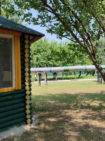 This media file shows an outdoor scene with a small wooden building in the foreground, a large pipe supported by structures in the middle ground, and trees and greenery in the background. The building is constructed of dark green painted logs with yellow trim around a window. The pipe is silver with green bands and is elevated above the ground. The setting appears to be a park or an area with some infrastructure. The sun is shining, casting shadows on the grass, suggesting it is daytime. The location is identified as Narowlya, Belarus. No people or text are visible in the image.