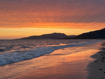 The image depicts a sunset scene on a beach in Tarifa, Spain. The sky is filled with dramatic orange and pink clouds, with a bright band of yellow light just above the horizon. Silhouetted against the colorful sky are rolling hills and mountains that form the coastline. The sea is calm, with gentle waves breaking on the shore. The wet sand on the beach reflects the colors of the sky, creating a mirror-like surface. Small stones and seaweed are scattered along the upper edge of the sand. No people or specific man-made structures are visible in the frame.