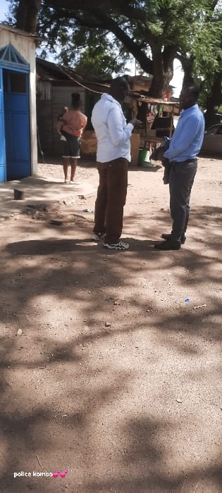 The image depicts an outdoor daytime scene in Homa Bay, Kenya. In the foreground, two adult males of dark complexion are standing face-to-face, appearing to engage in conversation on an unpaved dirt surface. The man on the left wears a light-colored long-sleeved collared shirt, brown trousers, and white athletic shoes; his right hand is raised slightly. The man on the right wears a light blue long-sleeved collared shirt, dark trousers, and black dress shoes; he holds a dark object in his left arm. In the background, to the left of the men, an adult female is visible, facing away from the camera, carrying a child secured on her back. She stands near a light-colored building with a blue door and a triangular decorative element above it. Behind the subjects, a mix of structures is visible, including makeshift dwellings with corrugated iron roofs and a small wooden stall displaying various items. Large trees with dense foliage cast shadows across the ground, which is composed of packed dirt and small debris. The environment suggests a semi-urban setting with a blend of natural elements and rudimentary construction.