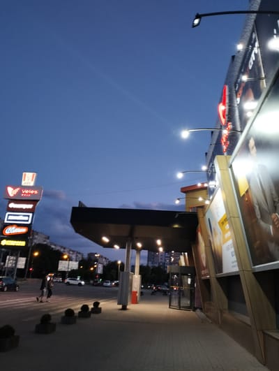 The image shows a street scene in Ivano-Frankivsk, Ukraine, at dusk. On the left, illuminated signs for various businesses, including KFC and Veles Mall, are stacked vertically. A couple is crossing the crosswalk toward the signs. Cars are visible on the street behind them. On the right, a modern building with large advertising panels is illuminated by several lights. A covered walkway extends from the building to the sidewalk. The sky is a gradient of blue, indicating the transition from day to night.