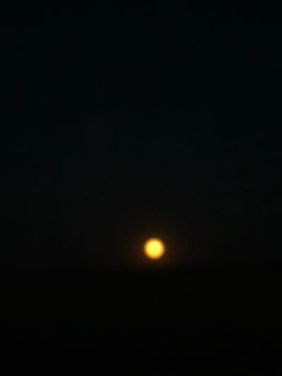 The image is a photograph of the night sky with a bright, out-of-focus celestial body, likely the moon or a planet, visible in the right half of the frame. The background is completely dark, suggesting it is nighttime. There is a subtle grain or noise present throughout the image. The celestial body emits a warm, yellowish-orange light and has a soft, diffused glow around it, indicating that it is not sharply in focus or that atmospheric conditions are affecting the light. The overall scene is stark and minimalist, with the single, luminous object being the sole point of interest against the vast darkness. No people, objects, text, or specific location cues are discernible beyond the dark sky. The provided location context of "Таганрогский, Russia" suggests a possible geographical origin for the photograph.