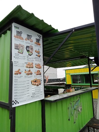 The image displays an outdoor food stall, likely a casual eatery, constructed from vibrant green corrugated metal. The sky is overcast, suggesting daytime with diffuse natural light.
The central feature is a large menu board attached to the side of the green stall, prominently displaying the brand name "BANGOR" at the top. The menu is divided into sections such as "PAKET FAMILY," "PAKET PARTY," and "SUPER HEMAT," showcasing various food items like fried chicken, burgers, and combo meals with accompanying images. Specific items visible include "SPICY CRAZY," "CHILL BOX," "PAKET SAMBORY," "Burger Bangor," and options like "Kentang" (fries) and "Nasi" (rice). Prices are clearly listed in Indonesian Rupiah (Rp), for example, "Rp 21.000" and "Rp 63.700." A black and white checkered pattern is incorporated into the menu design.
The stall has an open counter area where several condiment bottles, including ketchup and other sauces, are neatly arranged, suggesting readiness for service. The background behind the counter is dark, with equipment hinting at food preparation. No people are present, indicating a moment of quiet, perhaps before opening or after closing hours.
To the right and further back, another food stall, also made of corrugated metal but in yellow and green, is visible, featuring a window. The architectural style of both stalls, resembling modified shipping containers, suggests an informal, perhaps street-food, setting common in places like Kecamatan Panji, Indonesia. Stylized text, likely a brand or slogan, is embossed or painted on the lower green panel of the main stall, though not fully legible.