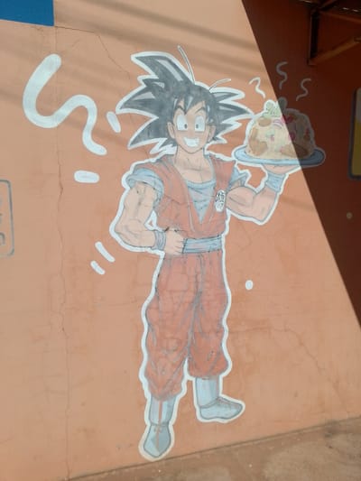 This is a close-up, eye-level, outdoor, full-shot photograph of a mural painted on an orange, cracked, stucco wall in São Gabriel Do Oeste, Brazil. The mural depicts the anime character Goku from Dragon Ball Z, holding a plate of food. Goku is rendered in a cartoonish style with spiky black hair, large eyes, a wide smile, and wearing his signature orange gi with blue undershirt and wristbands. He is also wearing blue shoes. The plate of food he holds appears to be a mound of rice and meat, topped with some green garnish and red onion slices, with steam rising from it. To the left of Goku, there is a stylized white question mark, and to the right, there are white wavy lines, suggesting steam or thought. The wall is textured and shows signs of age and wear, with visible cracks. A diagonal shadow, cast from an overhead structure, partially covers the right side of the mural and the wall. The top edge of the image shows a strip of blue painted surface above the orange wall.