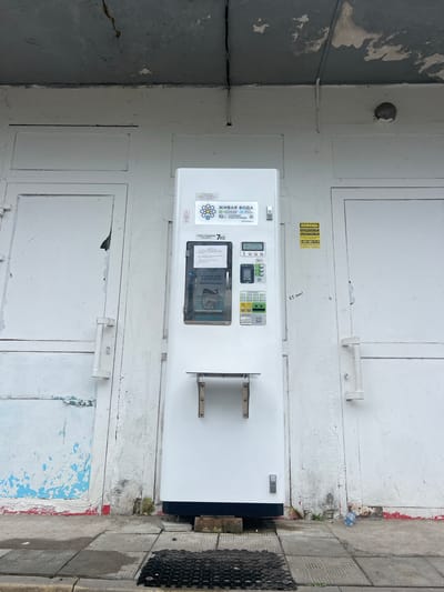 An outdoor view of a white water vending machine is centrally positioned in front of two white doors. The machine displays Russian text and has a digital screen, a keypad, and a dispenser area. Above the machine, the concrete ceiling shows signs of damage and cracking. To the right of the vending machine, a yellow sticker with Russian text and a phone number is affixed to the door. The ground in front of the machine is paved with square tiles and some debris. A plastic water bottle is visible on the ground to the right of the machine. The overall environment appears to be an outdoor urban setting, likely a commercial or public space in Kronstadt, Russia.