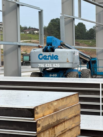 A blue and gray Genie aerial work platform is parked on a construction site in Stróża, Poland. The platform is positioned in front of a building under construction, characterized by its large gray metal beams. In the background, a green hill dotted with trees and a house can be seen. In the foreground, several large, flat, insulated panels are stacked, with some showing signs of rust. The sky is overcast. No people are visible in the image.