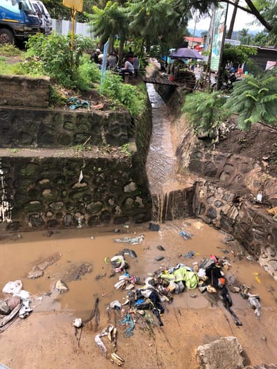 The image shows a flooded drainage ditch in Arusha, Tanzania, located at coordinates -3.363, 36.675. The ditch is lined with stone walls and filled with brown water. A wooden bridge spans the ditch, leading to a street beyond. The ditch is overflowing with trash, including plastic bags, clothing, and other debris. The water is murky and flowing quickly. There is a group of people seated near the edge of the ditch, but their actions are not visible. The surrounding environment is urban, with trees and buildings in the background.