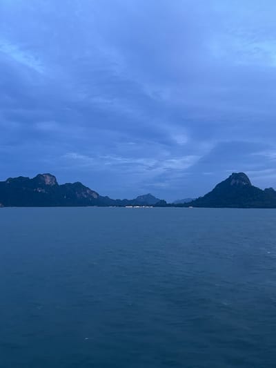 The image depicts a serene seascape in Don Sak, Thailand, during twilight. The sky, a deep, overcast blue, is filled with wispy clouds. In the distance, a series of islands with lush green vegetation and rocky outcrops emerge from the calm, dark blue water. The water's surface is gently rippled, reflecting the muted light. Small, distant lights are visible along the shoreline of one of the islands, suggesting a settlement. The overall atmosphere is tranquil and peaceful, with the fading light casting a cool, blue hue over the entire scene. There are no people or visible animals in the image.