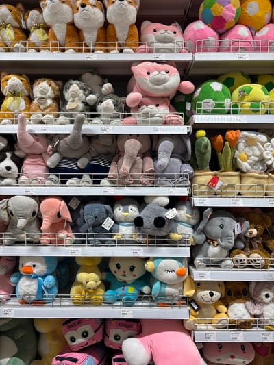 The image depicts a retail display of stuffed toys on shelves in a store. The toys include various animals such as cats, dogs, elephants, penguins, and rabbits, as well as plush balls and flower-shaped pillows. They are arranged in rows on white metal shelves, with some toys stacked on top of each other. Price tags are visible on some of the shelves. The overall environment appears to be a well-lit store with a focus on children's toys. The image was taken in Tangerang, Indonesia, within a retail environment. There are no people visible in the image, and the focus is solely on the merchandise.