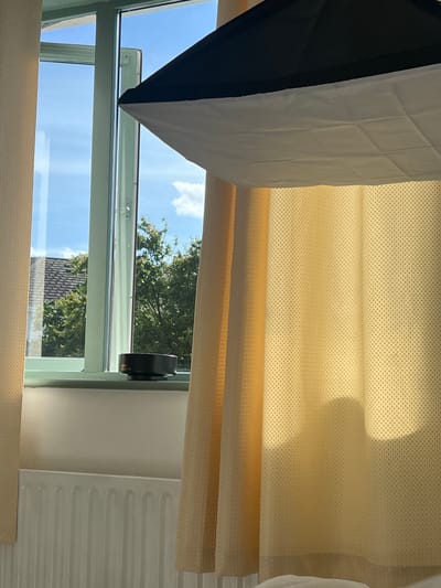 The image captures an indoor scene through a window with a light blue sky and scattered clouds visible outside. A window frame, painted light green, is prominent. On the windowsill, a black object, possibly a lens cap, rests to the left of center. To the right, a light yellow curtain with a subtle textured pattern is draped, obscuring part of the window and the wall. Above the window, a black and white photographic softbox is positioned, extending into the frame from the top. The lower portion of the image reveals a white radiator beneath the window. The scene appears to be set in Balbriggan, Ireland.