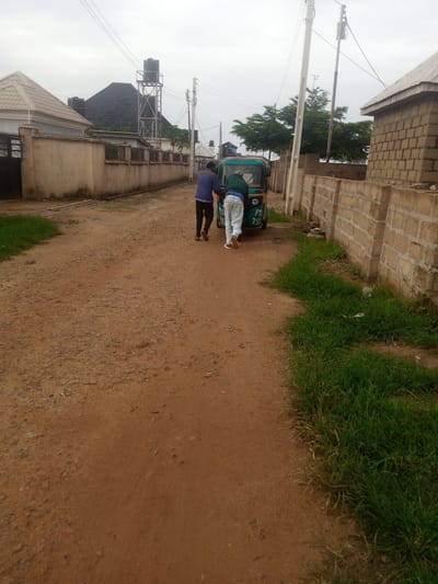 In Katon Rikkos, Nigeria, two individuals are seen pushing a teal and yellow motorized tricycle down a dirt road. The tricycle has "PTA 7910" written on its side. The men are dressed casually, one in a blue long-sleeved shirt and dark pants, the other in a green jacket and light-colored pants. They are positioned on the rear of the tricycle, appearing to push it forward. The environment is characterized by a dusty, unpaved road lined with a concrete wall on the right, constructed from light-colored blocks. Beyond the wall, more of the same block construction is visible. On the left side of the road, a beige wall with a metal gate encloses a property with a light-colored, peaked roof. Further down the road on the left, another building with a dark, steeply pitched roof is visible, as well as a water tower with a black tank. In the background, trees and power lines are present, indicating a somewhat rural or suburban setting within the city. The sky is overcast.