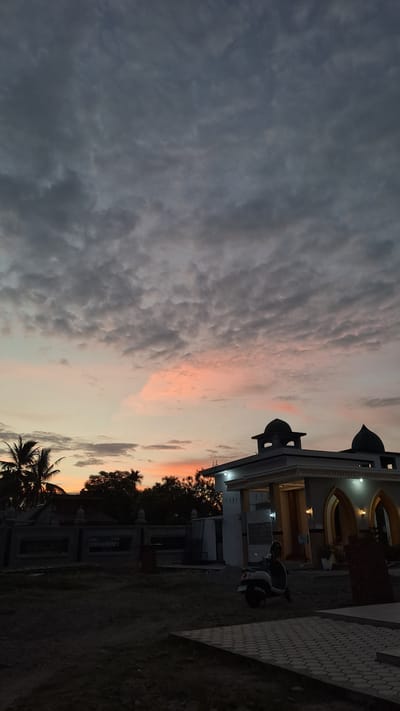 A mosque with two domes is visible against a cloudy sky at sunset. The sky has patches of pink and orange. In front of the mosque, a scooter is parked. Palm trees and other foliage are present in the background. A paved area is in the foreground. The scene appears to be in a city or town, possibly Lhokawe Teungoh, Indonesia.